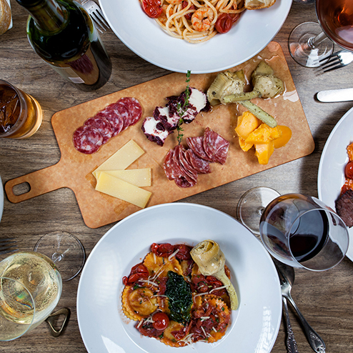 A table with plates of pasta, a charcuterie board with assorted meats and cheese, glasses of wine, and a bottle of wine.