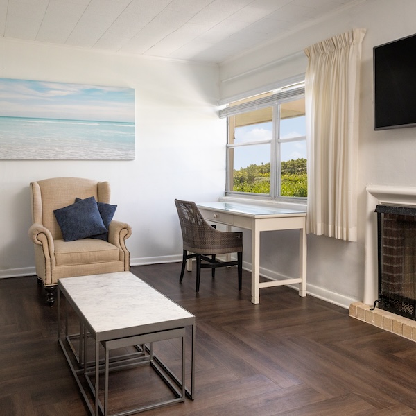 A room with a beige armchair, a desk with a chair, a wall-mounted TV, a coffee table, and a window with a green landscape view. A beach-themed painting is hung on the wall.