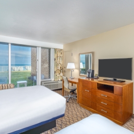 A hotel room with two beds, a TV on a dresser, a desk with a chair and lamp, and a sliding glass door with a view of the ocean.