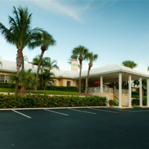 A low-rise hotel with a covered entrance flanked by palm trees, viewed from an empty parking lot during early evening.