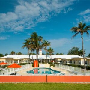 Tropical resort with a swimming pool surrounded by palm trees and low-rise white buildings under a clear blue sky.