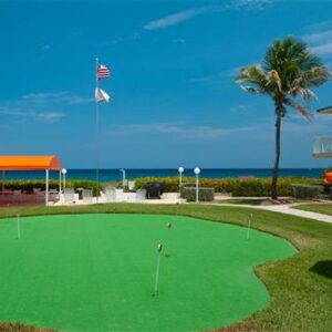 A vibrant oceanfront mini-golf course with palm trees, american flag, and bright orange umbrellas on a sunny day.