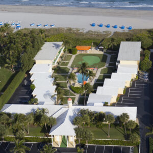 Aerial view of a coastal resort with symmetrical layout, featuring a central pool, palm trees, adjacent buildings, and a beach lined with blue umbrellas.