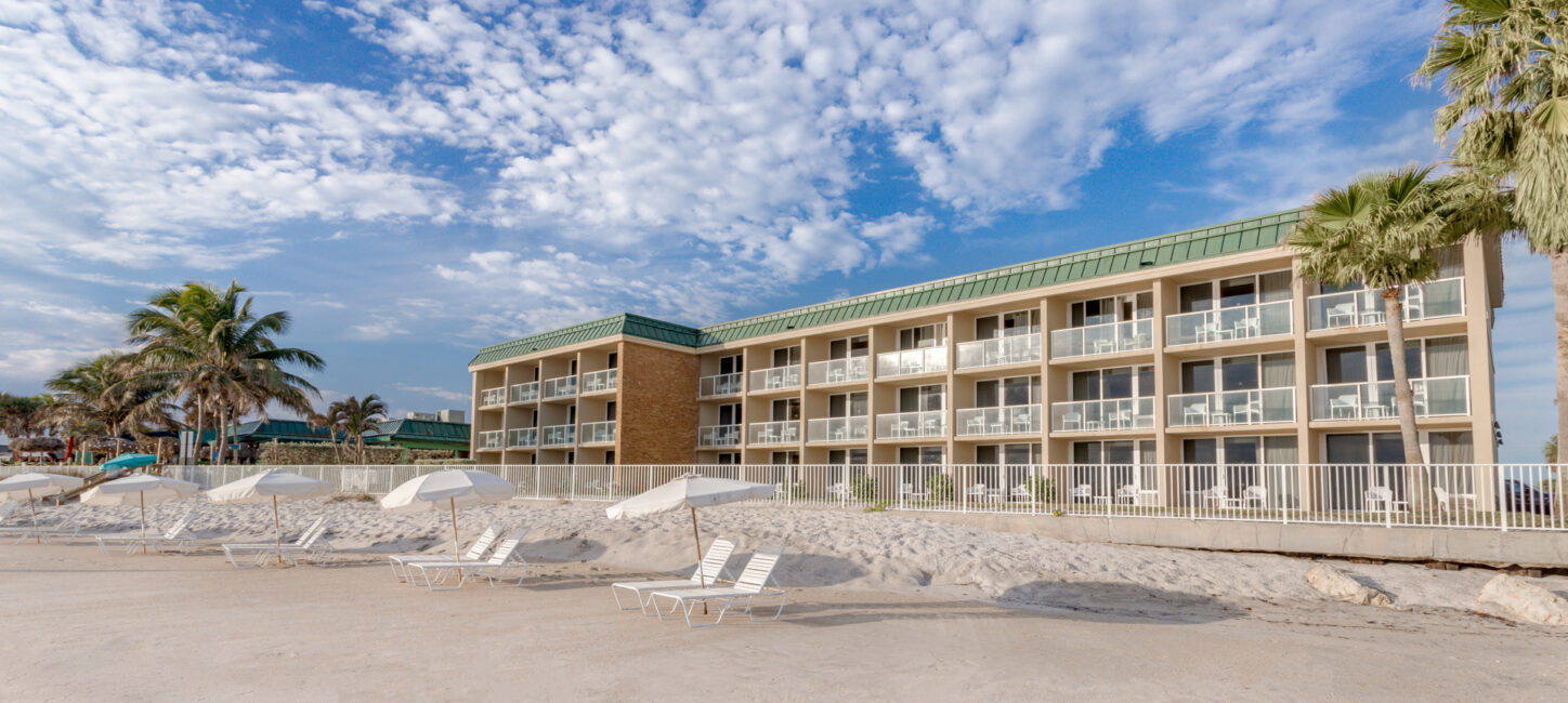 A beachfront hotel with multiple balconies under a cloudy sky, flanked by palm trees, with several beach chairs on the sand in the foreground.