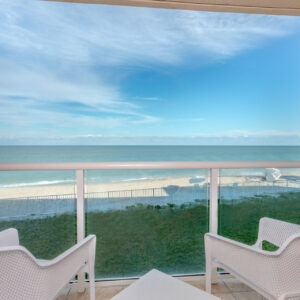 View from a balcony overlooking a beach with two chairs, a railing, palm tree to the left, and bright blue ocean and sky.