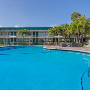 Outdoor swimming pool with lounge chairs and umbrellas in front of a two-story hotel on a sunny day.