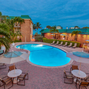 Twilight view of a hotel pool area with chairs, umbrellas, surrounded by palm trees and buildings under a blue evening sky.