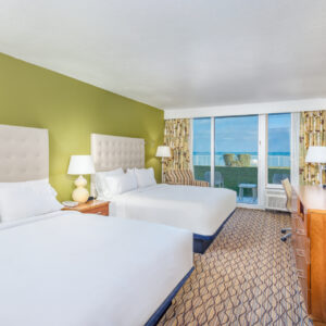 Bright hotel room with two beds, a desk, tv, and sliding doors opening to a balcony overlooking the sea.