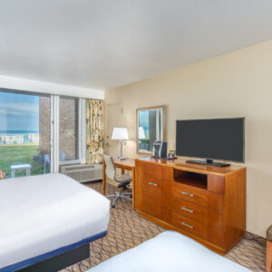 Hotel room with a bed, desk and chair, dresser with a large tv, and a window view of the ocean. bright and spacious interior.