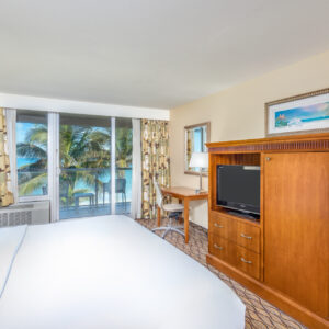 Bright hotel room with a large bed, wooden furniture, a desk, and a balcony view of palm trees.